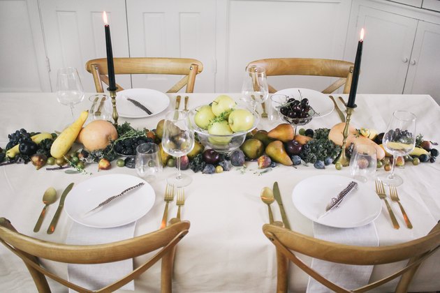 a line of produce, including grapes, pears, cherries, and squash runs down the length of a table with a white cloth and gold cutlery