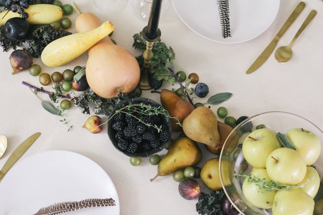 a line of produce, including grapes, pears, cherries, and squash runs down the length of a table with a white cloth and gold cutlery