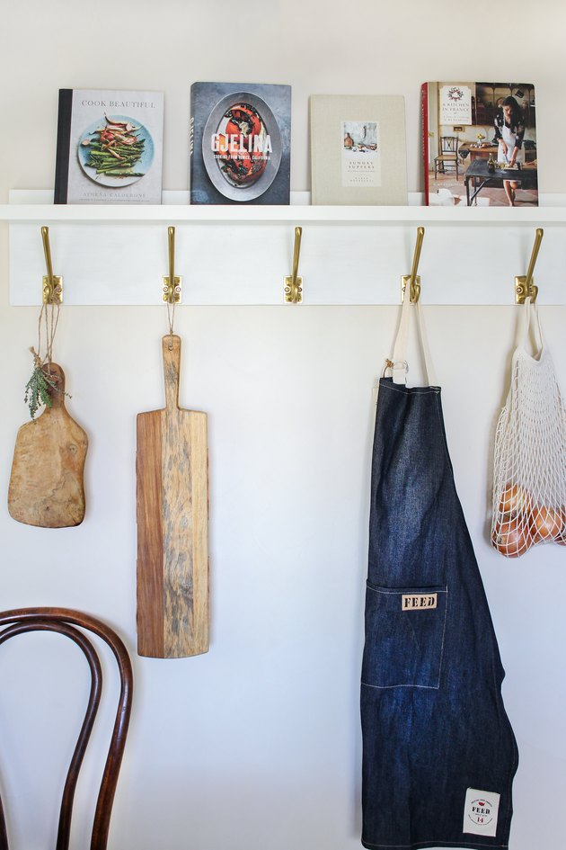 White kitchen wall hangers with gold hooks holding cutting boards, blue apron, and bag of tomatoes on white wall with books resting on top