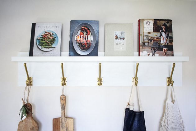 White kitchen wall hangers with gold hooks holding cutting boards, blue apron, and bag of tomatoes on white wall with books resting on top
