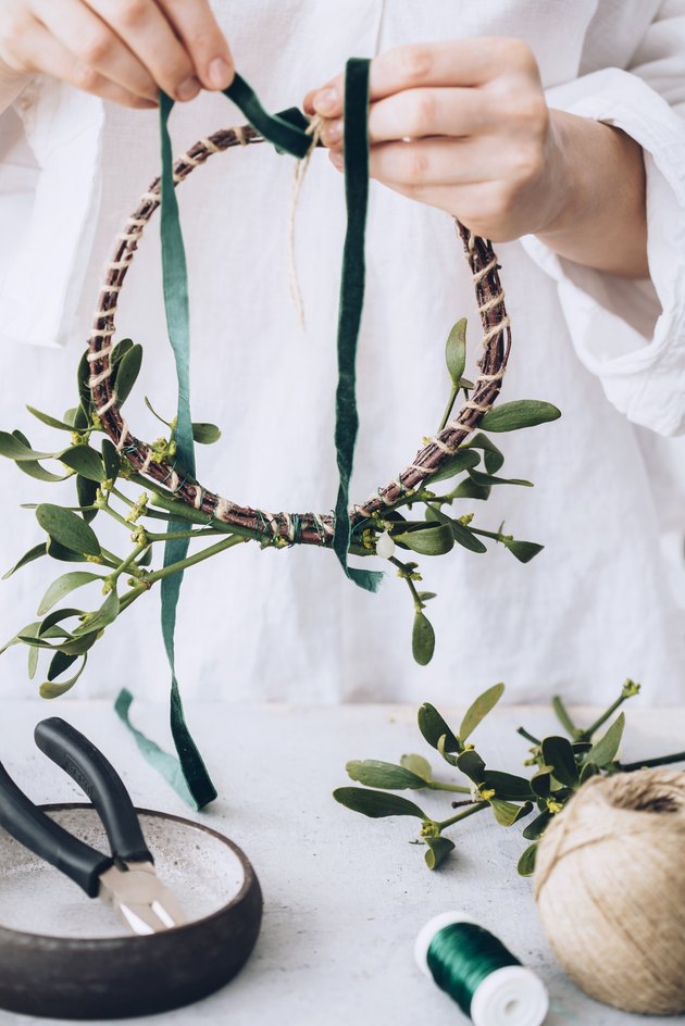 a woman wraps green ribbon around a diy mistletoe wreath in progress