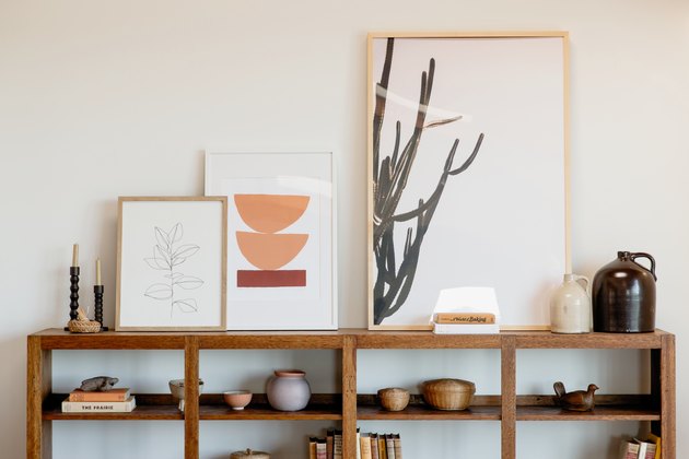 Wooden bookcase against a white wall. On its shelves, books and other decorative trinkets such as ceramic jugs, woven baskets, and a wooden dove figurine. On top, two candlestick holders and tree pieces of framed art.