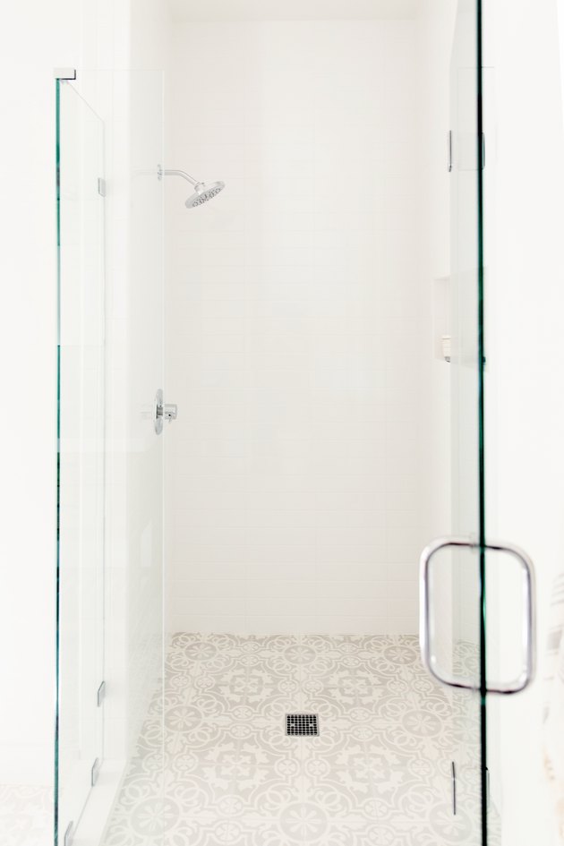 A glass shower door with a chrome handle in a white bathroom. Behind it, white shower with chrome fixtures and a white and grey patterned tile floor.