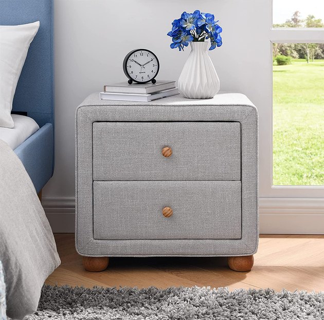 This new take on a nightstand is both practical and good-looking. It's crafted from wood and light gray linen for a warm addition to your space. Plus, its rounded legs add some extra detail and give the piece a high-end look.