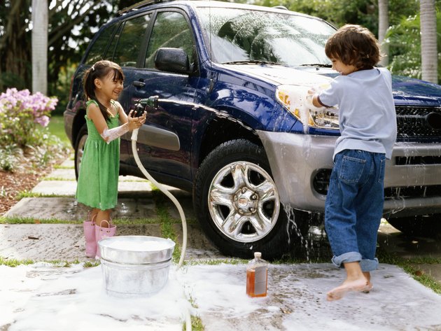 Girl (4-6) aiming hose at boy (5-7) washing car on driveway, smiling