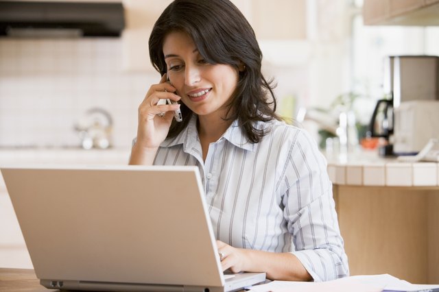 Woman in kitchen with laptop using cellular phone smiling