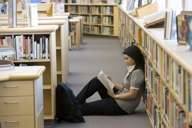 School girl (14-15) reading on floor by bookshelf in library