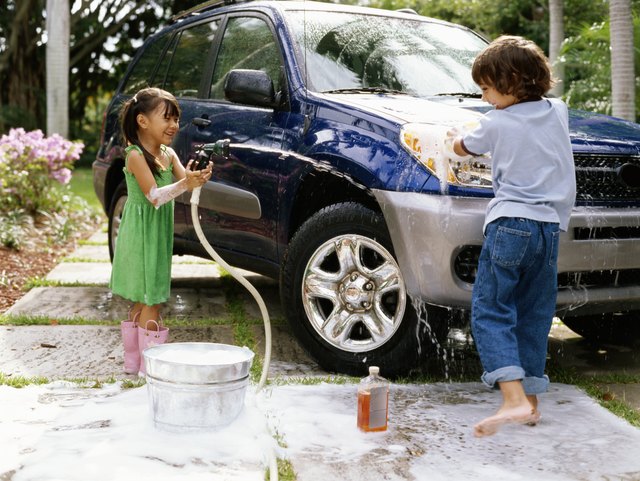 Girl (4-6) aiming hose at boy (5-7) washing car on driveway, smiling
