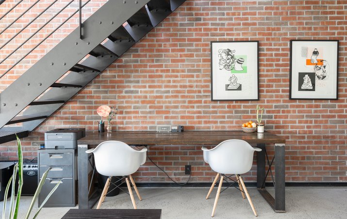 Black metal staircase against brick wall with wood table, white curved chairs, framed print, and small metal cabinet